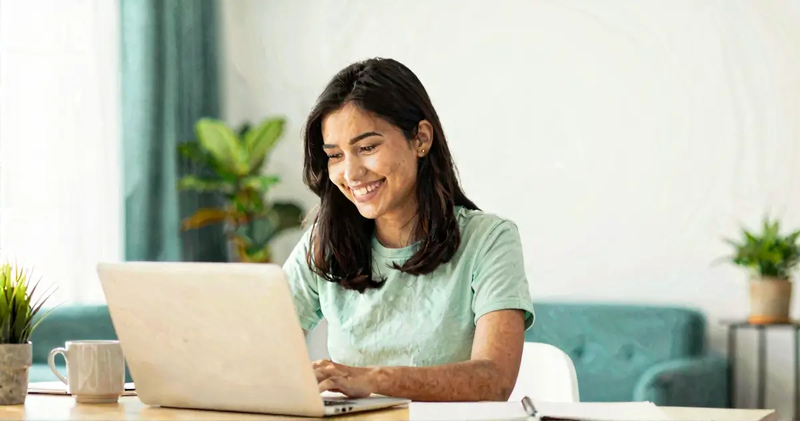 Smiling woman using a laptop at a desk in a bright room with indoor plants.